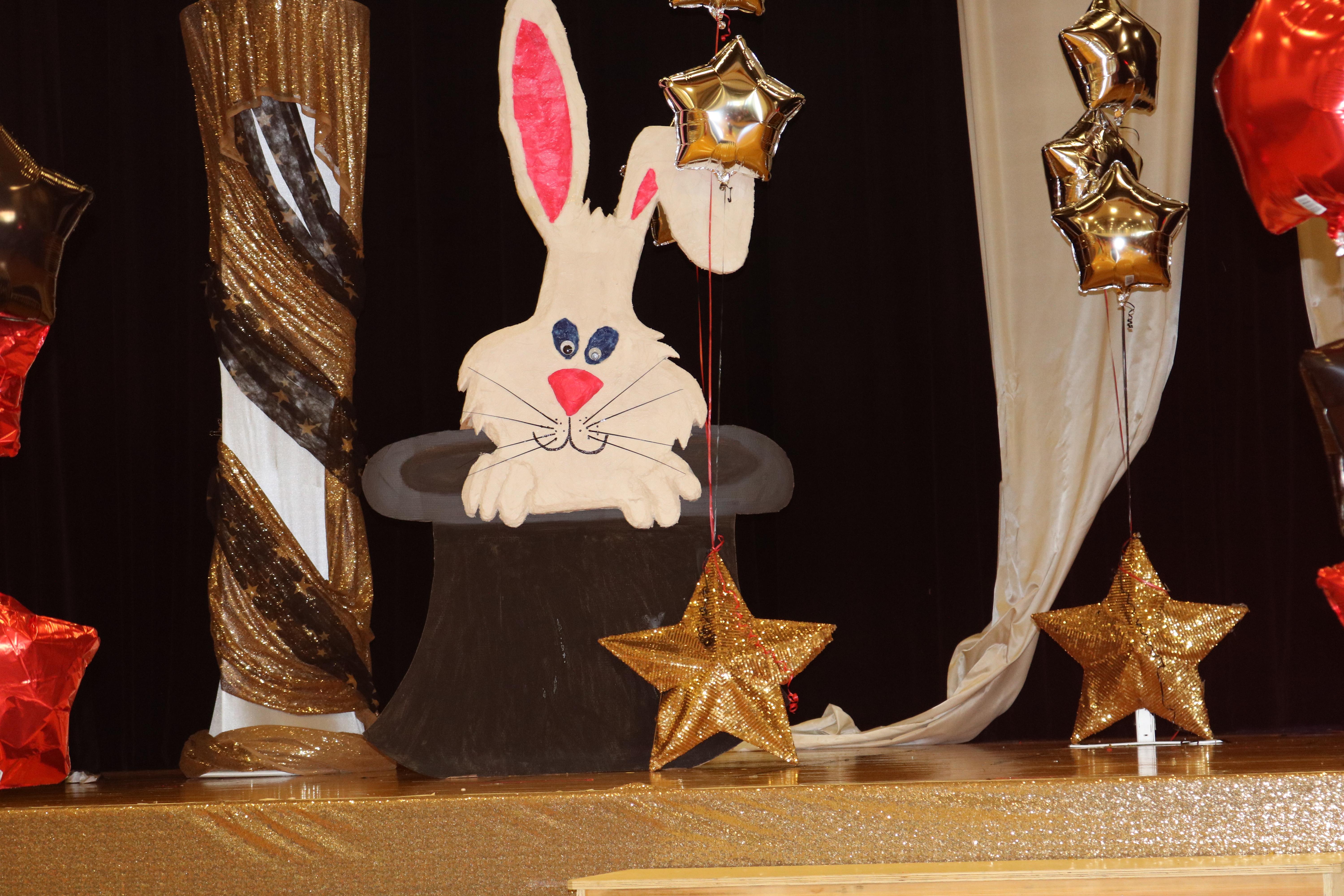 stage decorated with black hats and rabbits coming out with white and gold bunting and gold and red balloons